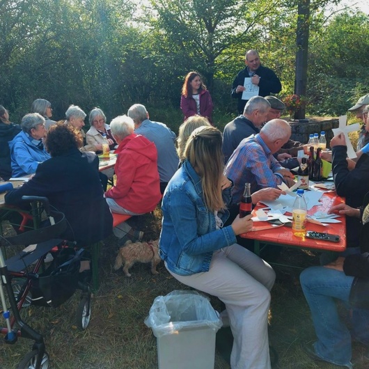 Blick auf eine Gruppe von Menschen. Sie sitzen an zwei Biertischen im Freien.